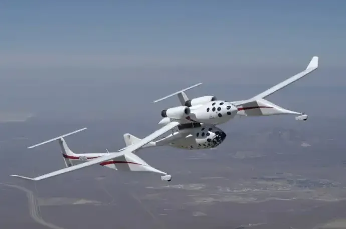A white plane with a unique double fuselage design flying in a clear blue sky over a desert landscape.