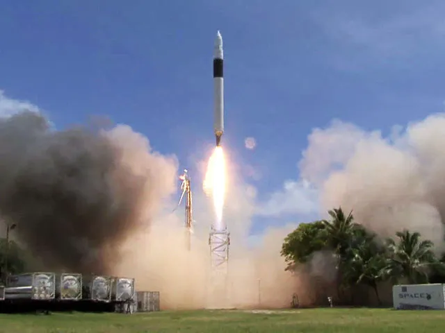 A rocket takes off into a clear blue sky with smoke billowing around the launch site.